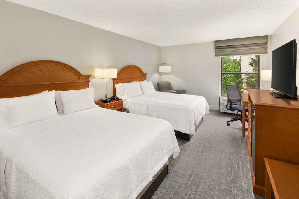 Two beds in a guest room with a chair, desk, TV, and mini-fridge at the Hampton by Hilton Inn